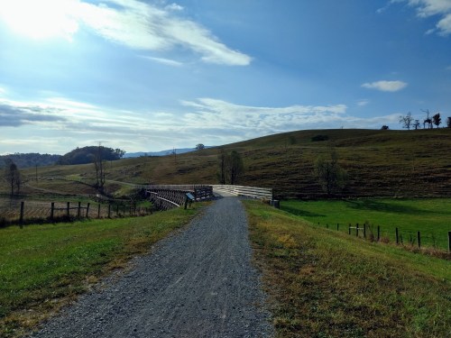 A gravel path leads to a wooden bridge, surrounded by rolling hills and a clear blue sky.