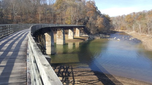 A wooden bridge spans a calm river, surrounded by trees and rocky banks under a clear blue sky.