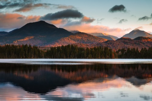 Serene lake reflecting colorful mountains and clouds at sunset, surrounded by lush forests.