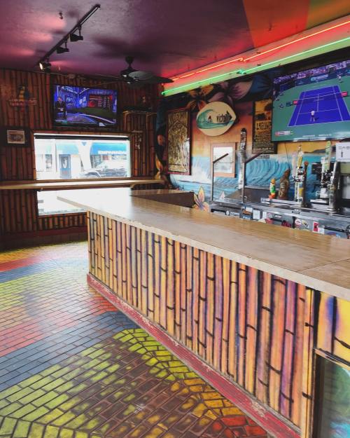 Colorful bar interior with bamboo decor, vibrant floor tiles, and TVs displaying sports. Brightly painted walls.