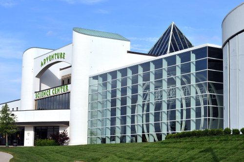 Modern science center with a glass facade and a triangular roof, surrounded by green grass and blue sky.