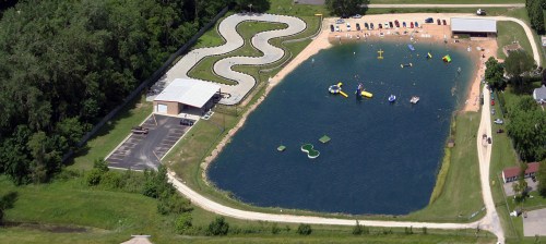 Aerial view of a lake with inflatable water toys, a sandy beach, and a winding path beside a building and parking area.
