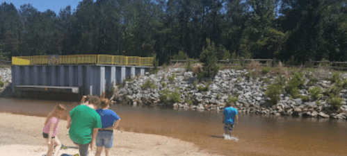 A group of people stands on a sandy bank by a shallow river, with a dam and trees in the background.