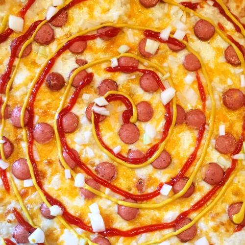 A close-up of a pizza topped with cheese, hot dog slices, onions, ketchup, and mustard arranged in a spiral pattern.