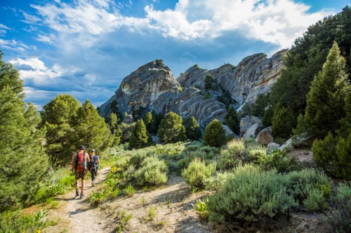 Two hikers walk along a trail surrounded by rocky formations and lush greenery under a cloudy sky.