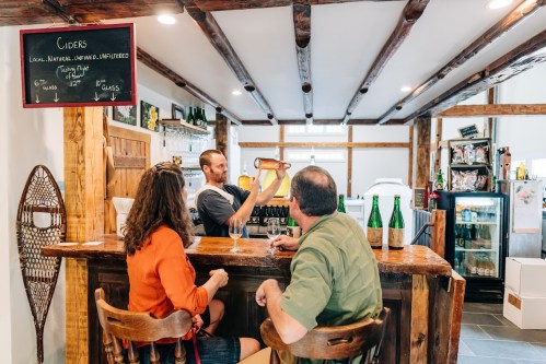A bartender serves cider to two customers at a rustic bar with wooden beams and shelves of bottles in the background.