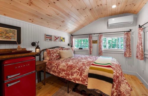 Cozy bedroom with wooden ceiling, floral bedding, red fridge, and large windows, featuring warm, inviting decor.