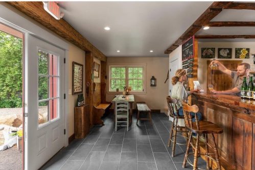A cozy bar area with a woman sitting at the counter and a bartender serving drinks, featuring rustic decor and wooden beams.