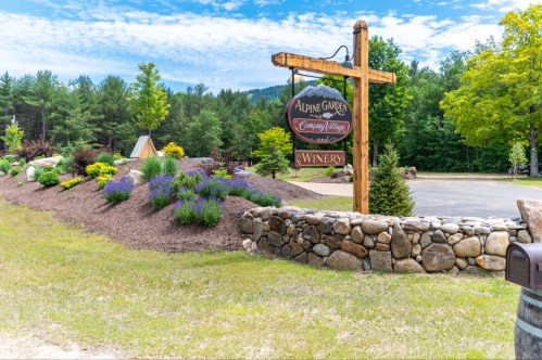 Sign for Alpine Garden Winery surrounded by colorful flowers and greenery, with a scenic backdrop of trees and mountains.