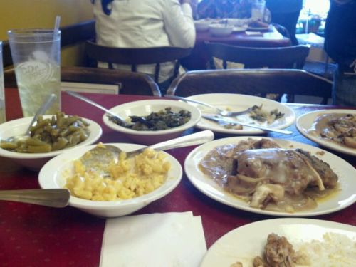 A table with several plates of Southern food, including greens, macaroni and cheese, and chicken with gravy, plus a drink.