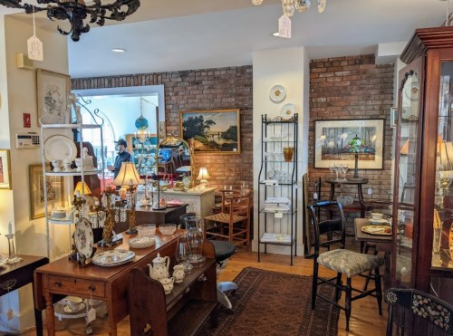 A cozy antique shop filled with vintage furniture, decor, and tableware, featuring warm lighting and brick walls.