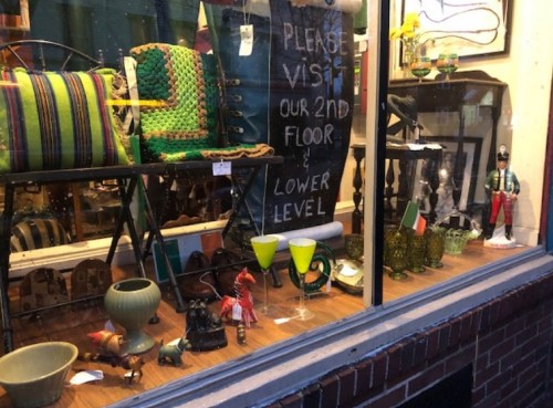 A shop window display featuring various antiques, decorative items, and a sign inviting visitors to explore further.