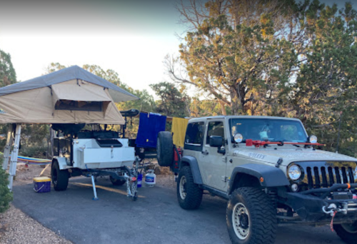 A Jeep with a trailer and rooftop tent set up in a wooded area, with clothes hanging on a line nearby.