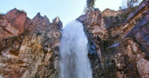 A powerful waterfall cascading down rocky cliffs, surrounded by rugged terrain and clear blue sky.