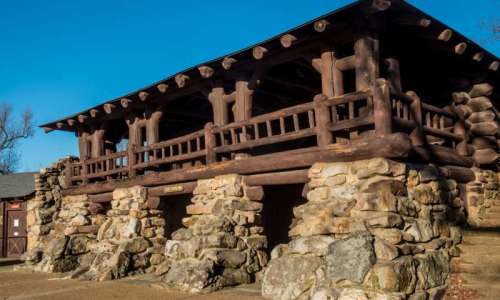 A rustic log cabin with stone foundations, featuring a balcony and surrounded by clear blue skies.