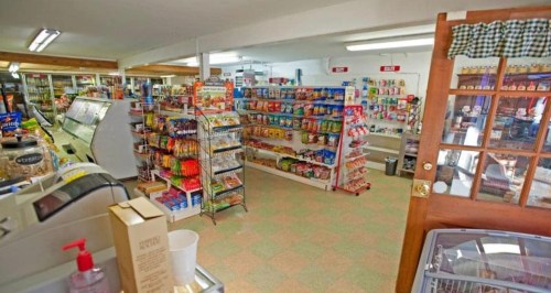 Interior of a small convenience store with shelves of snacks, drinks, and a checkout counter. Brightly lit and organized.