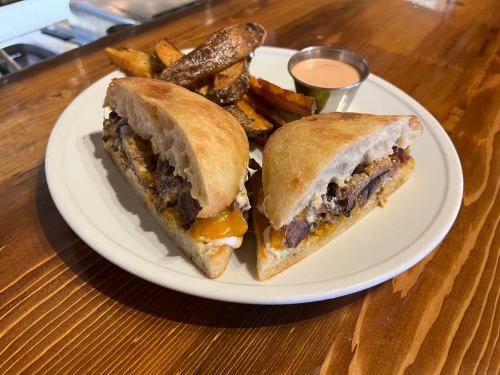 A plate with a sandwich filled with meat and cheese, served with crispy potato wedges and a small cup of dipping sauce.
