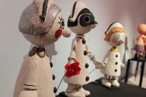 Three whimsical, painted wooden dolls in white outfits, with distinct facial features and accessories, displayed on a shelf.
