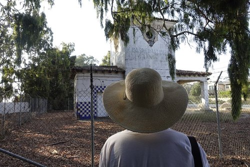 A person wearing a large straw hat stands in front of an old, abandoned building surrounded by trees and a fence.