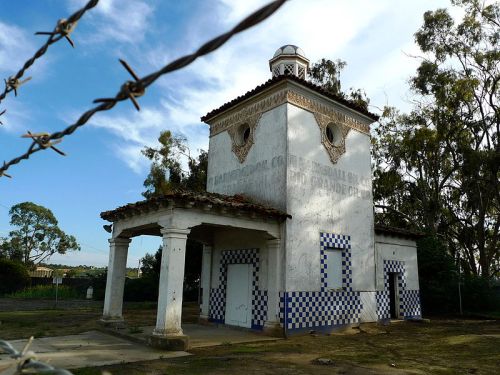 Abandoned building with a tower, featuring blue and white tiles, surrounded by trees and a barbed wire fence.