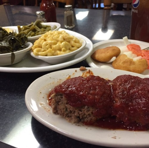 A plate of meatloaf with tomato sauce, served with macaroni and cheese, greens, cornbread, and a side of pickles.