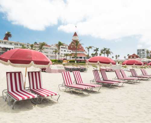 Beach scene with red and white striped lounge chairs and umbrellas, overlooking a historic hotel and palm trees.