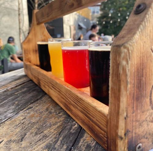 A wooden flight holder with four colorful beer samples on a rustic table, set outdoors.