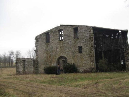 A weathered stone building with missing walls and windows, surrounded by overgrown grass and trees on a cloudy day.