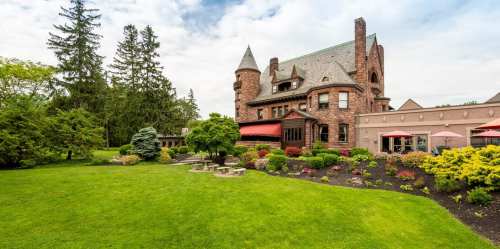 A large, historic mansion surrounded by lush green lawns and colorful flower beds under a cloudy sky.