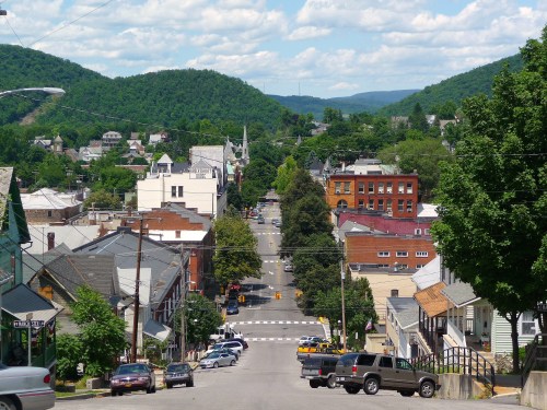 A scenic view of a small town with tree-lined streets, buildings, and mountains in the background under a blue sky.