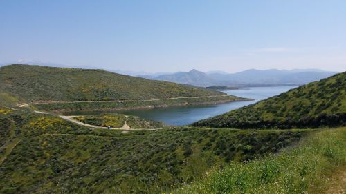 A serene landscape featuring a calm lake surrounded by green hills and distant mountains under a clear blue sky.