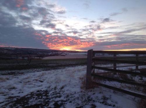 A winter landscape at sunset, featuring a wooden fence and colorful clouds over snow-covered fields.
