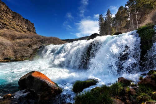 A cascading waterfall flows over rocks, surrounded by lush greenery and a clear blue sky.