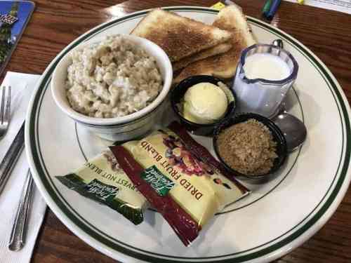 A plate with oatmeal, toast, butter, milk, dried fruit, and brown sugar on a wooden table.