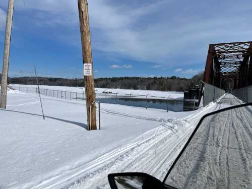 A snowy landscape with a frozen river and a metal bridge in the background, under a clear blue sky.