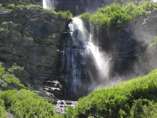A cascading waterfall flows down rocky cliffs, surrounded by lush green vegetation and mist rising from the water.