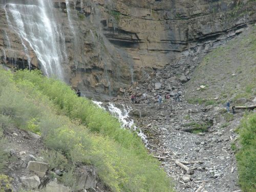 A waterfall cascades down rocky cliffs, surrounded by greenery and people exploring the rocky terrain below.