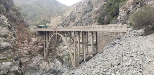 A concrete arch bridge spans a rocky canyon, surrounded by steep hills and greenery.