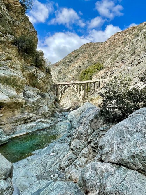 A scenic view of a rocky canyon with a bridge spanning across, under a blue sky with scattered clouds.