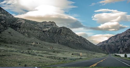 A scenic highway winds through mountains under a cloudy sky, with green hills and a distant house visible.