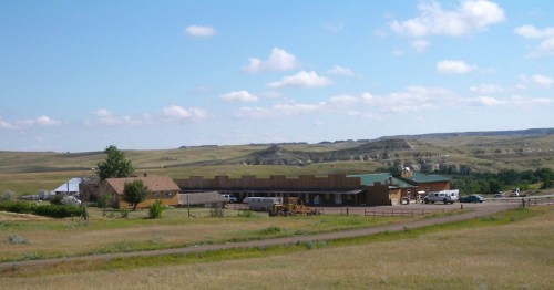 A rural landscape featuring a low building, vehicles, and rolling hills under a blue sky with scattered clouds.