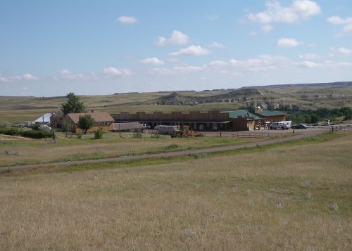A wide view of a ranch building surrounded by grassy fields and rolling hills under a partly cloudy sky.