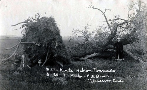 Two people stand beside a large uprooted tree after a tornado, with a damaged landscape in the background. Date: 5-26-11.