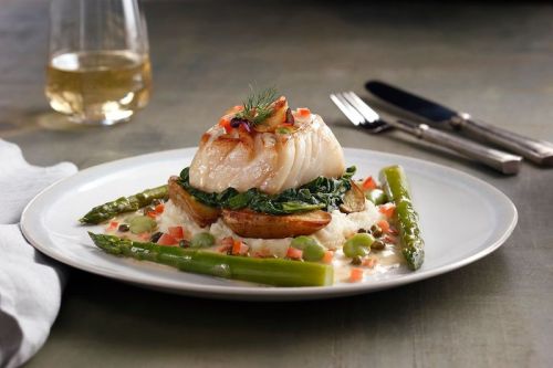 A plated dish featuring fish on a bed of greens, asparagus, and potatoes, garnished with herbs and colorful vegetables.