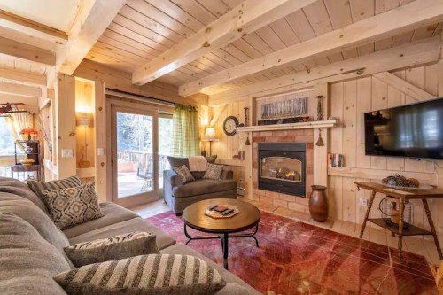 Cozy living room with wooden beams, a fireplace, comfortable seating, and large windows overlooking a deck.