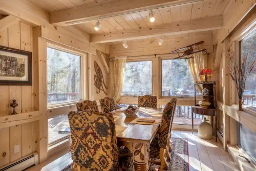 Cozy dining area with wooden walls, large windows, and patterned chairs, surrounded by natural light.