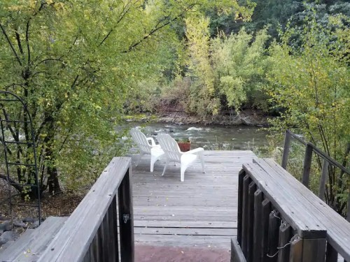 Two white chairs sit by a river, surrounded by lush green trees and autumn foliage, with a wooden deck in the foreground.
