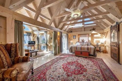 Cozy wooden cabin interior with a bed, patterned rug, and large windows, featuring rustic decor and a warm atmosphere.