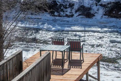 A wooden deck with a table and two chairs overlooking a frozen river and snowy landscape.