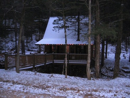 A cozy cabin with a metal roof, surrounded by snow-covered trees and a wooden deck, illuminated by festive lights.